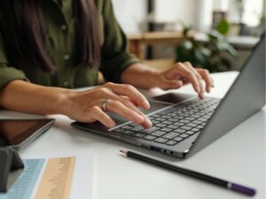 Hand of young female teacher or student typing on laptop keyboard while sitting by desk in front of camera and answering task questions